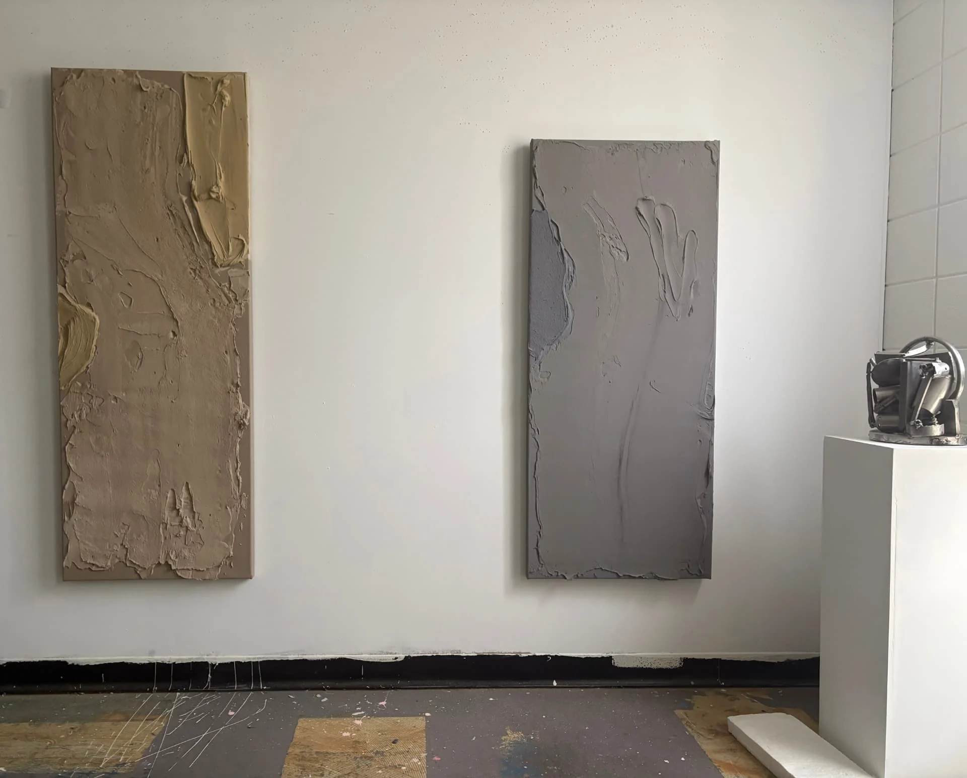 Two textured paintings hung on a studio wall beside a welded steel sculpture on a plinth